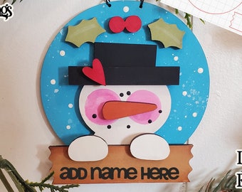 May include: A wooden snowman ornament with a blue background, white dots, and a red heart on the hat. The snowman has a pink face, black eyes, a carrot nose, and a brown sign that says "add name here".