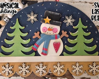 May include: A wooden snowman-themed wall hanging. The snowman has a black top hat, a blue scarf, and a red heart. The background is dark blue with snowflakes and green trees. The text "WHIMSICAL Inkings" and "SVG LASER READY" are visible.