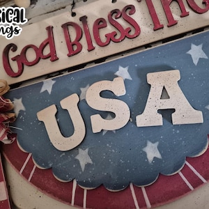 May include: Wooden patriotic sign with the text "God Bless The USA". The sign features a blue and red scalloped design with white stars and the letters "USA". A bow and the text "Whimsical Inklings" are also visible.