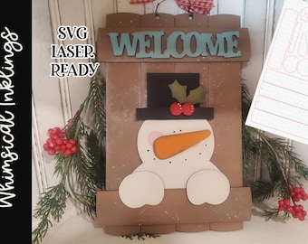 May include: A wooden welcome sign with a snowman face and a blue welcome sign. The sign is decorated with a red bow and holly. The sign is laser cut and ready to be assembled.