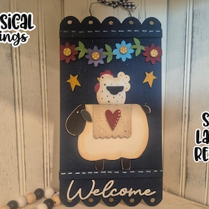 Peut inclure: Panneau de bienvenue en bois bleu marine avec un mouton, un cœur, des fleurs et des étoiles. Le panneau porte le texte "Welcome" en bas et "Whimsical Inklings" en haut. Le panneau est prêt pour le laser.