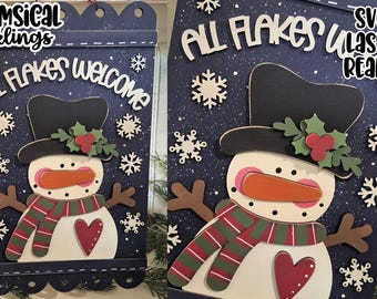May include: A wooden snowman sign with the text "ALL FLAKES WELCOME". The snowman has a black top hat, an orange carrot nose, and a red heart. The sign is decorated with snowflakes and a green and red scarf.