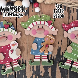 May include: Three whimsical wooden Christmas elf ornaments. Each elf has a unique design, including a snowman, gingerbread man, and candy cane. The ornaments are painted in festive colors with the text "Whimsical Inklings" and "SVG Laser Ready".