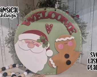 May include: A round, wooden Christmas sign with a green background and the word "WELCOME" in red. Santa Claus and a gingerbread man are featured. The sign also includes the text "WHIMSICAL Inkling" and "SVG LASER READY".