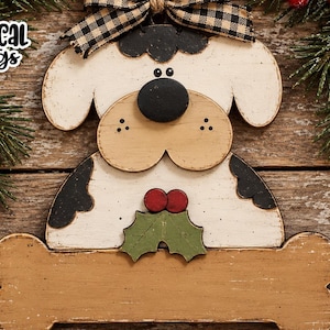 May include: A wooden dog-shaped sign with a tan bone-shaped base, featuring a white and tan dog with black ears and a black nose. The dog is adorned with a green holly leaf and two red berries. A black and tan buffalo check bow sits on top. Text reads Whimsical Inklings and SVG Laser Ready.