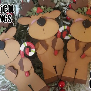 May include: Three wooden reindeer ornaments with candy canes in their hands. Each reindeer has a red nose, brown antlers, and a green holly wreath around its neck. The ornaments are laser cut and ready to be assembled.  SVG Laser Ready.