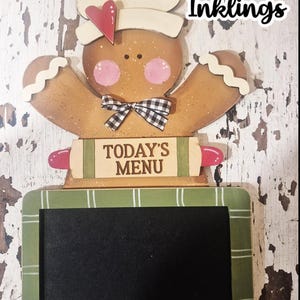 Op de afbeelding: Houten gingerbread man-vormig schoolbord met een koksmuts en een strikje. Het schoolbord heeft een groen-witte geruite rand en de woorden "TODAY'S MENU". De tekst "WHIMSICAL Inklings" is ook inbegrepen.