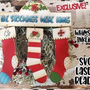 May include: A wooden Christmas stocking hanger with the text "The Stockings Were Hung" and three red and white striped stockings with different embellishments. The stockings are hanging from the sign. The sign is decorated with a red flower and green leaves. The background is a rustic wooden surface.