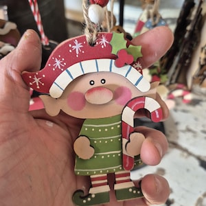 May include: Wooden Christmas elf ornament with a red and white hat, green dress, and candy cane. The elf has rosy cheeks and is holding a candy cane. The ornament is hanging from a twine string, perfect for holiday decor.