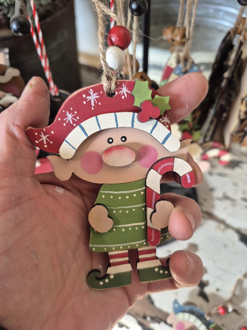 May include: Wooden Christmas elf ornament with a red and white hat, green dress, and candy cane. The elf has rosy cheeks and is holding a candy cane. The ornament is hanging from a twine string, perfect for holiday decor.