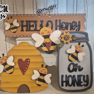 Könnte beinhalten: Ein Set aus vier Holzschildern mit Bienen-Motiv und den Texten "Hello Honey" und "Oh Honey". Die Schilder sind in Gelb, Braun, Grau und Schwarz mit weißen Akzenten bemalt. Die Schilder sind perfekt für einen Landhausstil oder eine rustikale Dekoration.