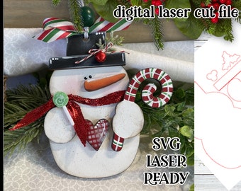May include: A white wooden snowman ornament with a red scarf, a candy cane, and a red and white heart. The ornament is surrounded by greenery and has a digital laser cut file for crafting.