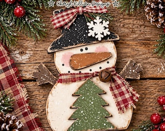 Puede incluir: Un adorno de madera con forma de muñeco de nieve que incluye un sombrero negro con un copo de nieve blanco, una nariz de zanahoria marrón y un árbol de Navidad verde. Lleva una bufanda de cuadros rojos y blancos con un cascabel y un lazo.