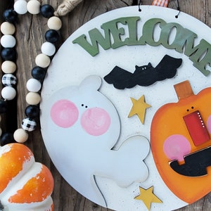 Pode incluir: Uma placa redonda de madeira branca com tema de Halloween com a mensagem "WELCOME" em verde, um morcego preto, um fantasma, uma abóbora e estrelas amarelas. A placa inclui o texto "SVG LASER READY" e "Whimsical Inklings".