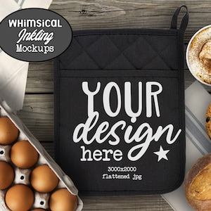 May include: Black quilted oven mitt with white text that says "YOUR design here" and a small star. The oven mitt is on a wooden surface with a carton of eggs, a cup of coffee, a cookie, and a loaf of bread.