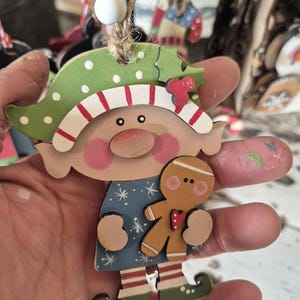 May include: Wooden Christmas ornament in the shape of a whimsical elf holding a gingerbread man. The elf has a green hat with white polka dots and red and white striped trim. The elf's dress is blue with white snowflakes.