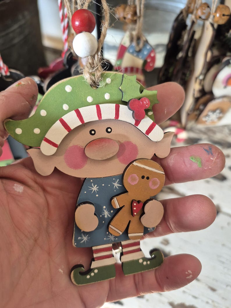 May include: Wooden Christmas ornament in the shape of a whimsical elf holding a gingerbread man. The elf has a green hat with white polka dots and red and white striped trim. The elf's dress is blue with white snowflakes.