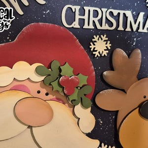May include: A wooden Christmas decoration featuring Santa Claus and a reindeer, with the word "CHRISTMAS" in large letters. The design includes snowflakes and the text "SVG Laser Ready" and "Whimsical Inklings".