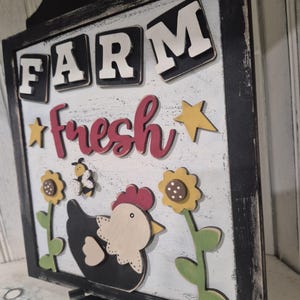 Peut inclure: Panneau de style ferme avec les mots "FARM fresh" en lettres blanches et rouges. Le panneau représente une poule noire et blanche, des tournesols et une abeille. Le panneau est encadré en noir et a un aspect vieilli.