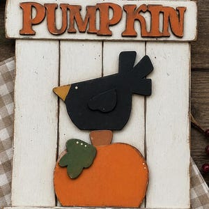 May include: A white wooden sign with the text PUMPKIN PATCH, featuring a black bird perched on an orange pumpkin with a green leaf. The sign includes orange and green lettering, two yellow stars, and an orange checkered ribbon at the top.