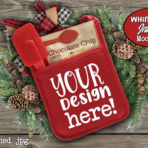 Op de afbeelding: Een rode ovenwant met een zak en een spatel. De zak heeft een wit ontwerp met de tekst "YOUR design here!" en de tekst "Chocolate Chip Cookie Mix" is zichtbaar op de verpakking in de zak. De ovenwant is omringd door een groene dennentak met dennenappels.