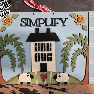 May include: A blue and white wooden sign with the word "SIMPLIFY" in black letters. The sign features a house with windows, two trees, and two sheep. The sign is decorated with sunflowers and a red heart.