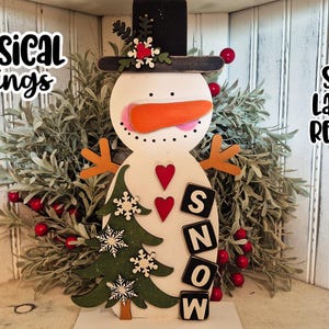May include: A wooden snowman decoration with an orange carrot nose, black top hat, and orange stick arms. The snowman has red heart accents and the word "SNOW" spelled out with black and white letter blocks. The text "Whimsical Inklings" and "SVG Laser Ready" are also visible.