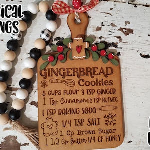 Pode incluir: Um ornamento de madeira em forma de receita de biscoitos de gengibre com um design de azevinho vermelho e verde. O ornamento é marrom com texto branco que diz "Gingerbread Cookies" e inclui os ingredientes para os biscoitos de gengibre. O texto diz: "3 xícaras de farinha, 3 colheres de chá de gengibre, 1 colher de chá de canela, 1/2 colher de chá de noz-moscada, 1 colher de chá de bicarbonato de sódio, 1/4 colher de chá de sal, 1 xícara de açúcar mascavo, 1 1/2 xícara de manteiga, 1/4 xícara de mel". O ornamento está pendurado em uma fita xadrez vermelha e branca.