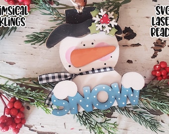 May include: A wooden snowman ornament with a black hat, a red nose, and a blue "SNOW" sign. The ornament is decorated with white polka dots and is surrounded by green pine branches and red berries.