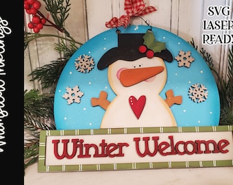 May include: A wooden snowman door hanger with a blue background and snowflakes. The snowman has a black hat, orange nose, and red heart. The sign reads "Winter Welcome".