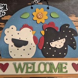 Puede incluir: Un letrero de bienvenida de madera con dos gallinas estilizadas sobre un fondo azul, con un girasol y hojas verdes. La palabra "WELCOME" está escrita debajo, con un corazón a cada lado. El letrero está listo para corte láser.