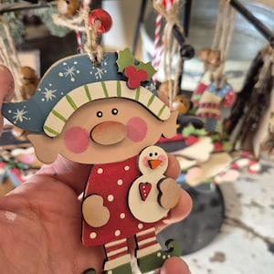 May include: Wooden Christmas ornament featuring a whimsical elf design. The elf wears a blue hat with white snowflakes and a red polka-dot dress. The elf holds a snowman. The ornament is strung with twine.