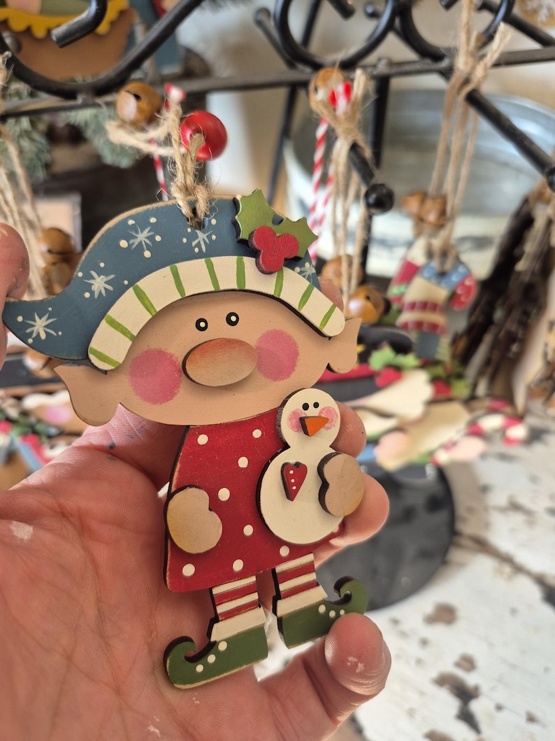 May include: Wooden Christmas ornament featuring a whimsical elf design. The elf wears a blue hat with white snowflakes and a red polka-dot dress. The elf holds a snowman. The ornament is strung with twine.