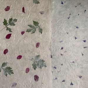 May include: Two pages of handmade paper with pressed flowers and leaves. One side features red petals and green leaves, while the other side has purple and blue flowers.