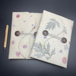 May include: Two handmade journals with decorative pressed flower covers. The journals are bound with twine and have wooden buttons. The covers are a cream color with pink and blue accents.