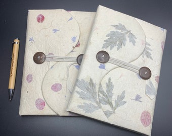 Peut inclure: Deux journaux faits à la main avec des couvertures décoratives de fleurs pressées. Les journaux sont reliés avec de la ficelle et ont des boutons en bois. Les couvertures sont de couleur crème avec des accents roses et bleus.