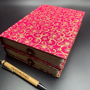 May include: Two pink and gold journals with a floral pattern on the cover. The journals are stacked on top of each other and have a white, fluffy binding. A bamboo pen with the words "Love Every Page" engraved on it is lying on a black surface.