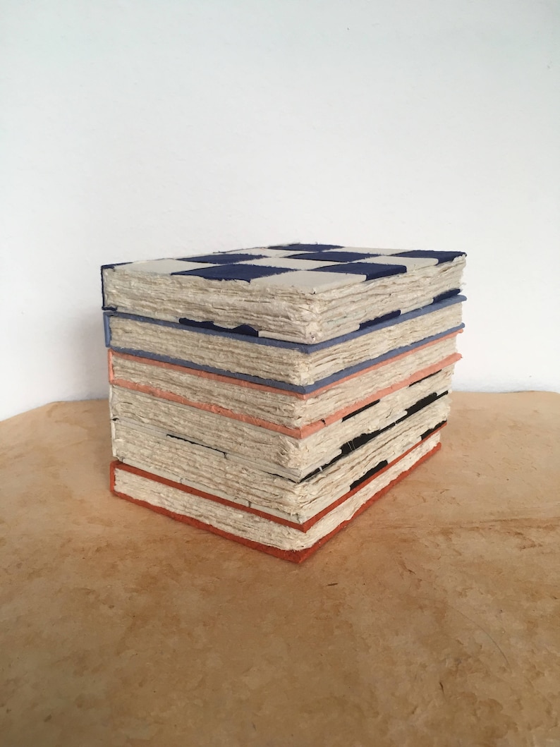 K&ouml;nnte beinhalten: Ein Stapel handgemachter B&uuml;cher mit einem Schachbrettmuster auf dem obersten Buch. Die B&uuml;cher sind mit einem hellblauen, orangefarbenen und schwarzen Besatz gebunden. Die B&uuml;cher liegen auf einer braunen Oberfl&auml;che.