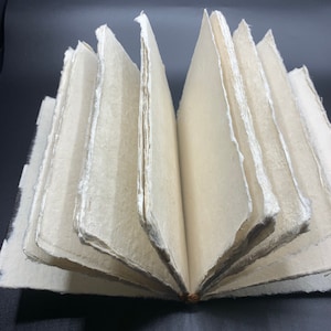 K&ouml;nnte beinhalten: Ein handgemachtes Journal mit einem nat&uuml;rlichen, cremefarbenen Papierumschlag. Die Seiten sind mit einer einfachen Naht gebunden und haben einen rauen, strukturierten Rand. Das Journal ist ge&ouml;ffnet, um die leeren Seiten im Inneren zu zeigen.