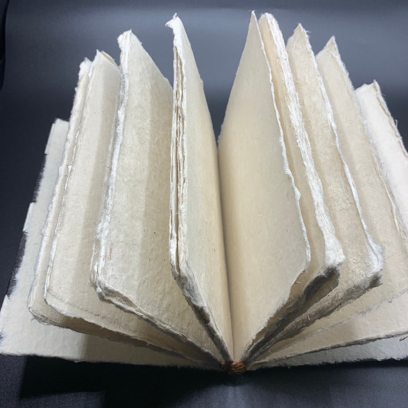 K&ouml;nnte beinhalten: Ein handgemachtes Journal mit einem nat&uuml;rlichen, cremefarbenen Papierumschlag. Die Seiten sind mit einer einfachen Naht gebunden und haben einen rauen, strukturierten Rand. Das Journal ist ge&ouml;ffnet, um die leeren Seiten im Inneren zu zeigen.