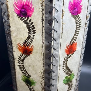 May include: A handmade paper lantern with a rectangular shape. The lantern features a floral design with pink, orange, and green flowers. The edges are stitched with black thread, and a red tassel hangs from the bottom. The lantern is a decorative item.