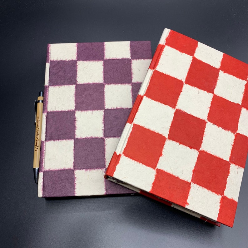 K&ouml;nnte beinhalten: Zwei karierte Notizb&uuml;cher, eines mit violetten Quadraten und eines mit roten Quadraten, auf einer schwarzen Oberfl&auml;che. Ein Holzstift mit dem Text "Home Sweet Home" liegt neben den Notizb&uuml;chern.