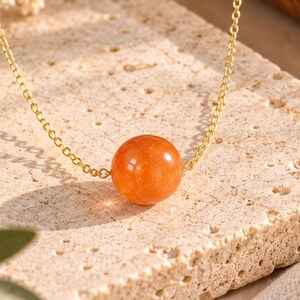 May include: A delicate gold chain necklace featuring a single, smooth, spherical orange gemstone bead as the central pendant. The chain is thin and metallic, holding the polished, translucent orange stone in a simple, minimalist arrangement.