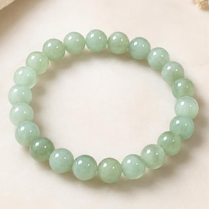 May include: A circular stretch bracelet composed of smooth, polished, pale green spherical beads. The beads are uniform in size and arranged in a continuous loop, showcasing a translucent, light mint-green color with subtle variations in tone.