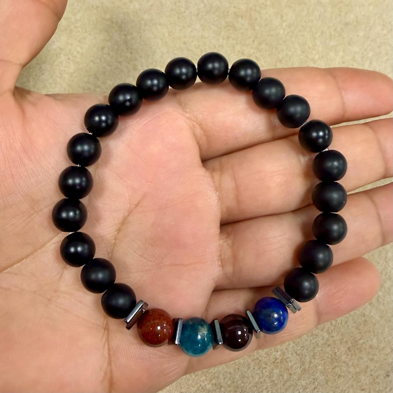 May include: A beaded bracelet featuring a series of matte black spherical beads interspersed with small metallic spacers. The design includes four distinct accent beads in shades of reddish-brown, teal, dark maroon, and deep blue near the bottom.