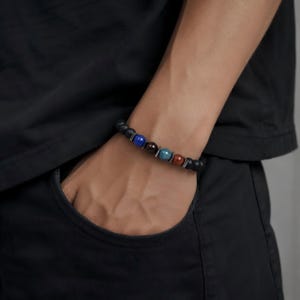 May include: A beaded bracelet featuring matte black spherical beads interspersed with four polished accent beads in blue, dark brown, teal, and reddish-brown. The bracelet includes small metallic spacers between the colored beads.