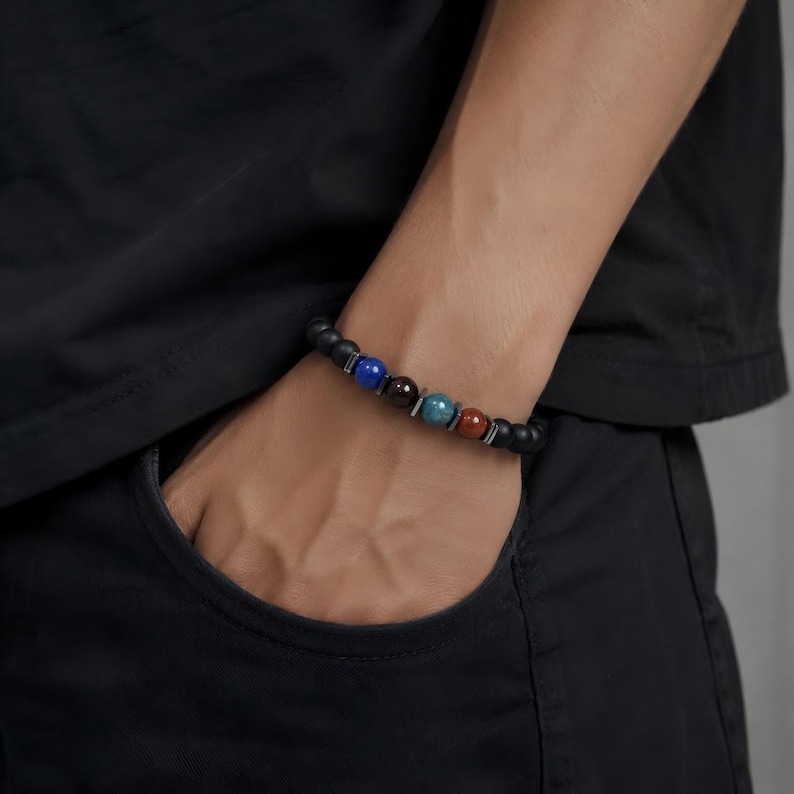 May include: A beaded bracelet featuring matte black spherical beads interspersed with four polished accent beads in blue, dark brown, teal, and reddish-brown. The bracelet includes small metallic spacers between the colored beads.