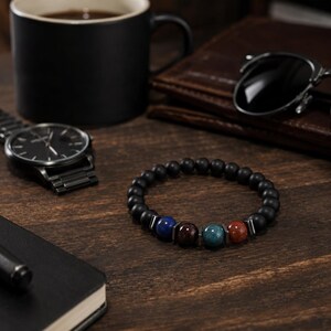 May include: A beaded bracelet featuring matte black spherical beads interspersed with four polished, multi-colored stone beads in blue, dark brown, teal, and reddish-brown, separated by small, flat, metallic-toned rectangular spacers.