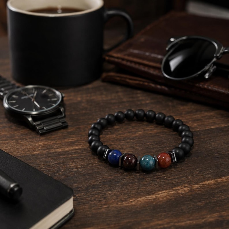 May include: A beaded bracelet featuring matte black spherical beads interspersed with four polished, multi-colored stone beads in blue, dark brown, teal, and reddish-brown, separated by small, flat, metallic-toned rectangular spacers.