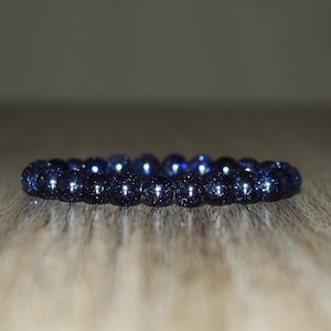May include: A dark blue beaded bracelet with a sparkling, speckled appearance. The beads are round and evenly sized, strung together to form a complete circle. The bracelet is displayed on a wooden surface.